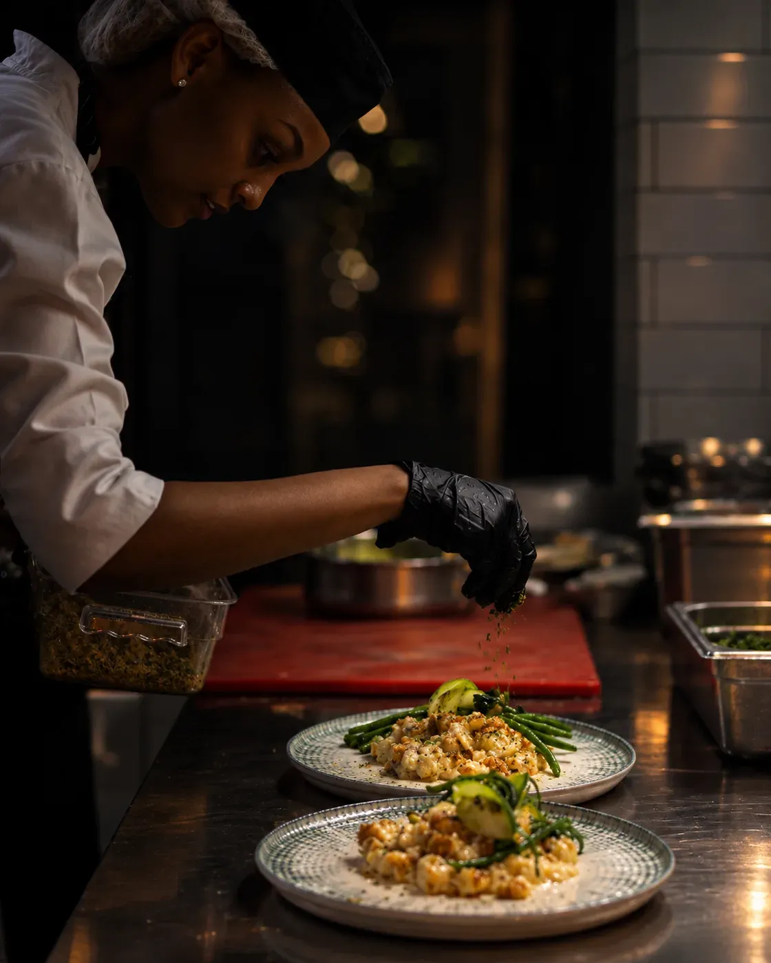A chef sprinkling herbs over two plated dishes at the pass
